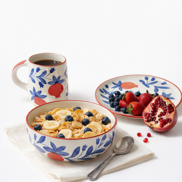 A coordinated breakfast table setting with a floral ceramic bowl of cereal, a matching mug, a plate with fruit, and a spoon on a white background. The dishes have a white base with blue leaf motifs and red/orange accents, and the scene includes strawberries, blueberries, a pomegranate, and cereal.