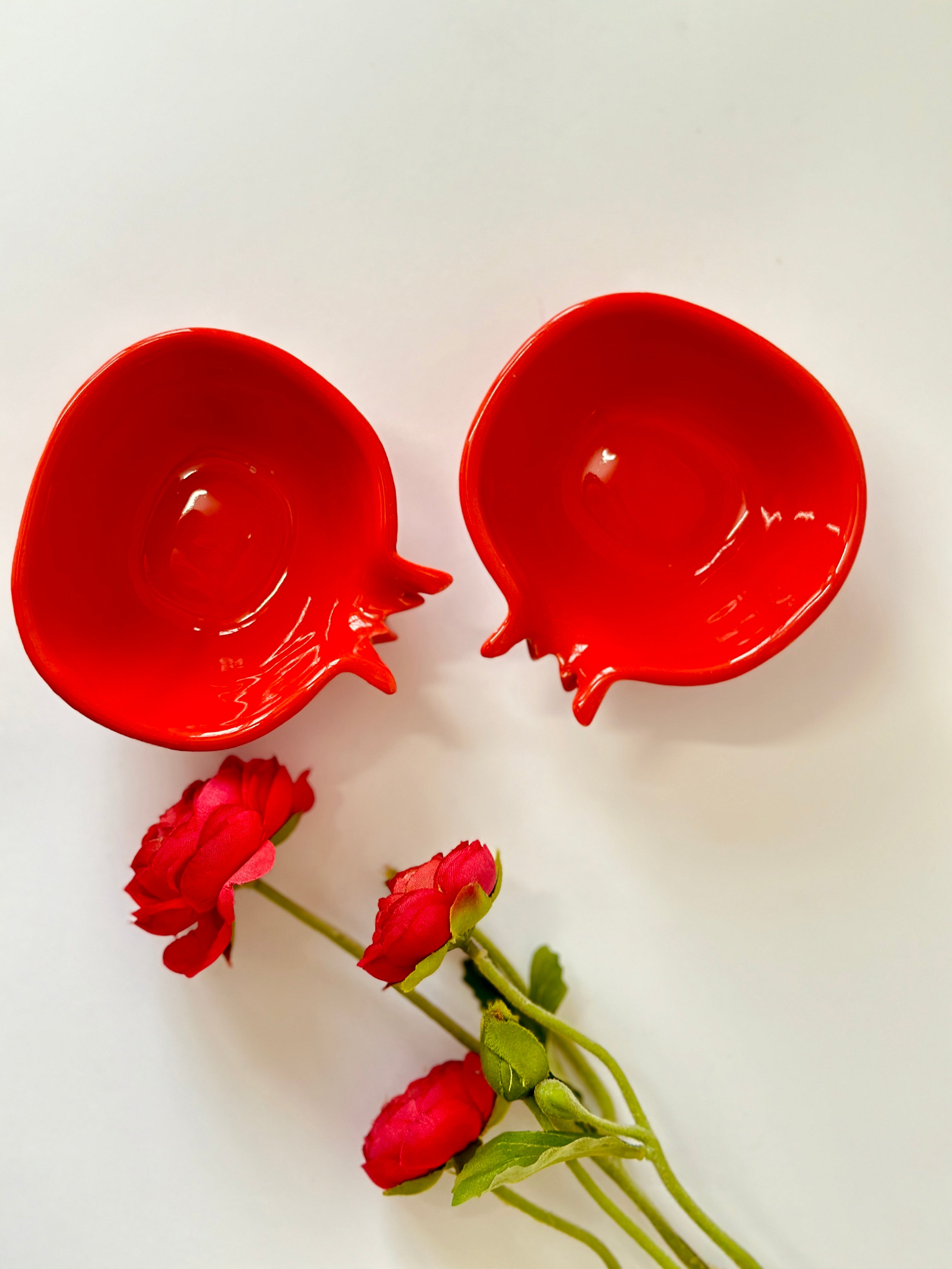 Two red ceramic chip dip bowls with a pomegranate fruit design, accompanied by two red roses on a white background.