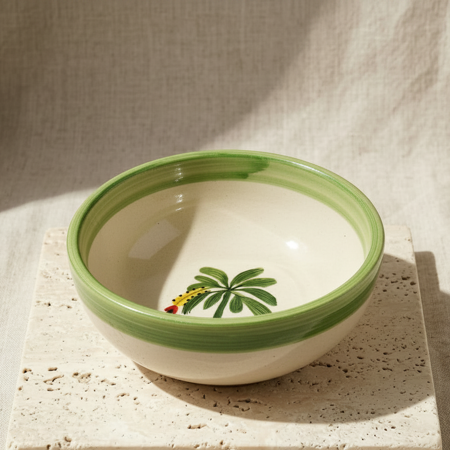 A hand-painted ceramic bowl with a green rim and a small green palm leaf design inside, resting on a square beige stone coaster against a textured beige linen background.