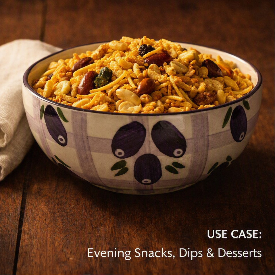 A ceramic bowl filled with a crunchy snack mix of nuts, seeds, and dried fruit, set on a dark wooden table next to a folded beige cloth. The bowl has a white base with a purple plaid pattern and purple fruit motifs.