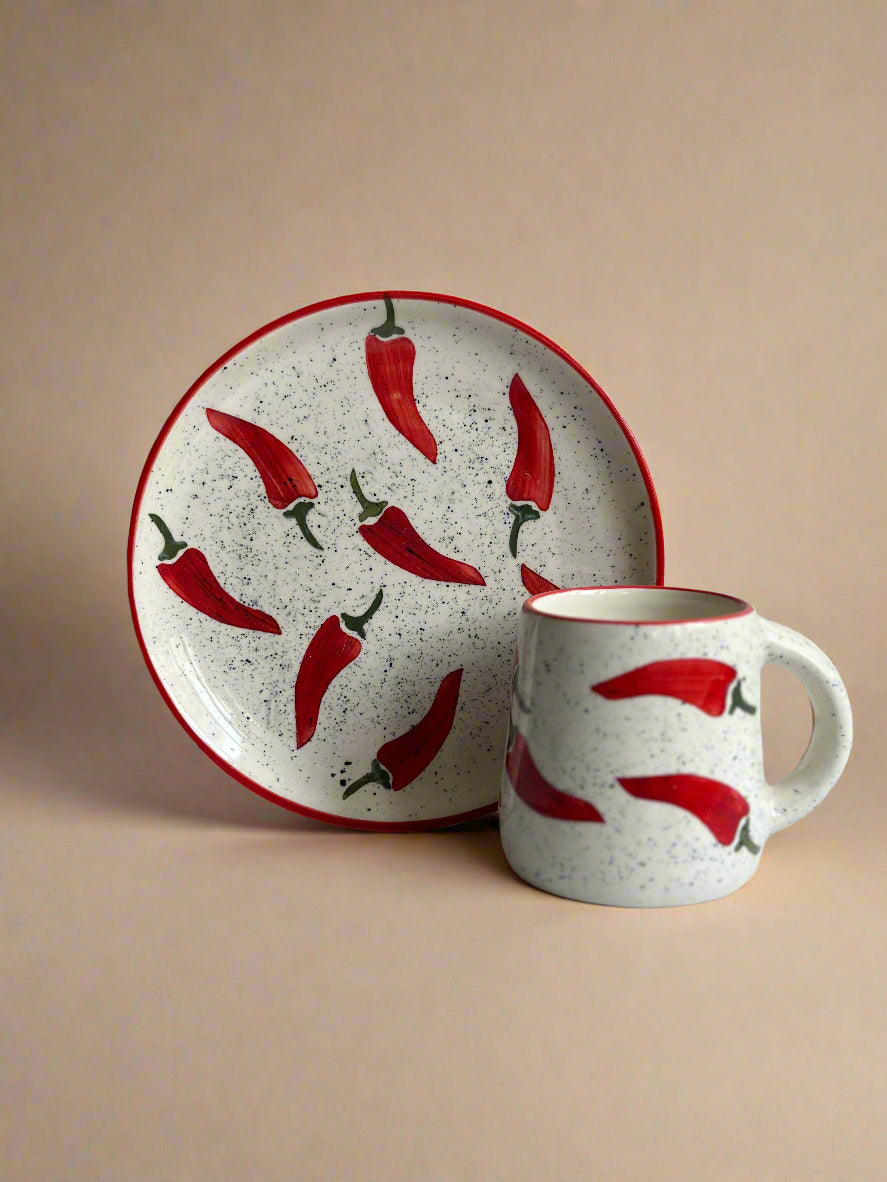 Ceramic mug and plate with red chili pepper design on a white surface.