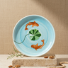 A round ceramic plate with a light blue background, decorated with two orange fish and a green four-leaf clover, displayed on a stone block with decorative props.