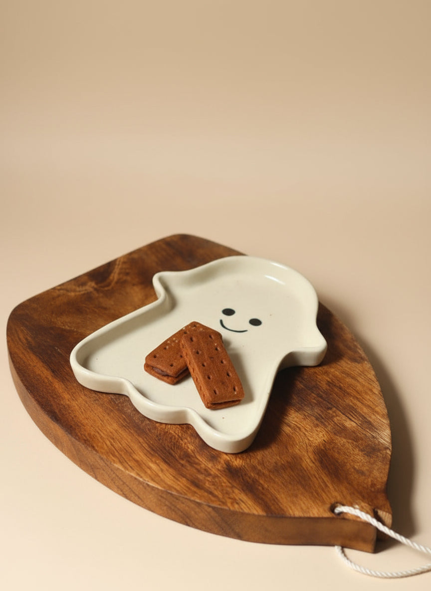 White ghost-shaped plate with a smiley face holding a cookie on a wooden board against a beige background