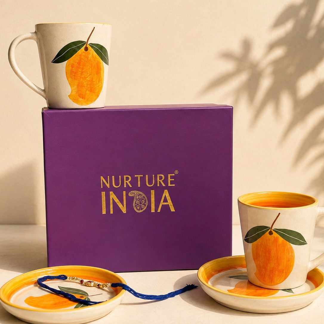 Two white ceramic mugs with orange mango illustrations, displayed with matching saucers and a purple Nurture India gift box on a beige surface with warm natural shadows.