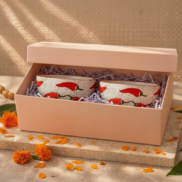 A pair of white ceramic tea cups with red chili pepper prints, presented inside a light pink gift box with purple shredded paper. The box is styled on a stone slab with orange marigold flowers and green leaves against a textured beige background.