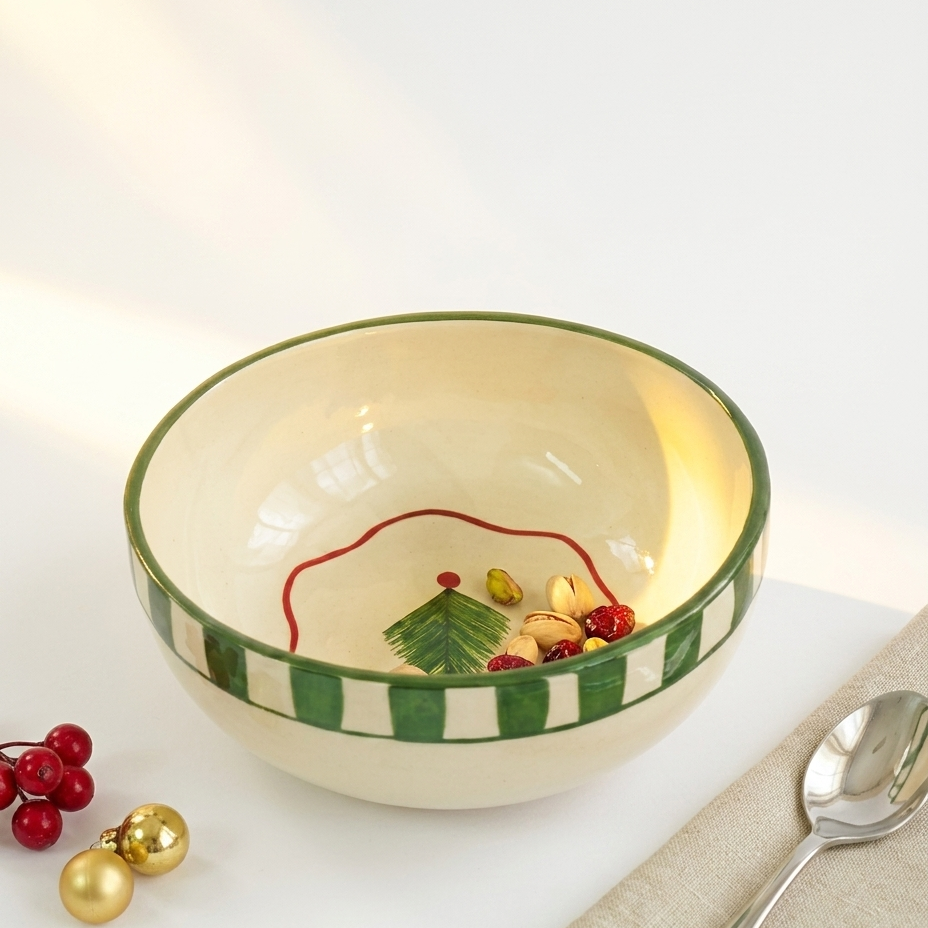 A ceramic Christmas bowl with a cream-colored interior, a green and white striped rim, and a painted green tree with red berries inside. The bowl is styled on a white surface with a beige napkin, a silver spoon, and red and gold holiday decorations.