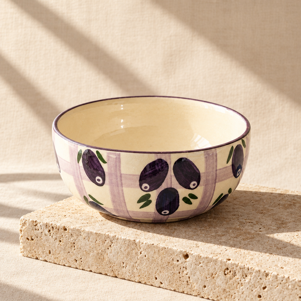 A ceramic bowl with a cream background, purple and lavender plaid pattern, and painted purple olives with green leaves, resting on a folded teal cloth on a gray stone surface.
