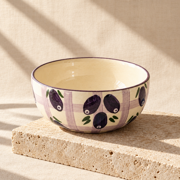 A ceramic bowl with a cream background, purple and lavender plaid pattern, and painted purple olives with green leaves, resting on a folded teal cloth on a gray stone surface.
