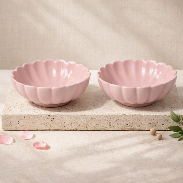 Two pink scalloped ceramic bowls displayed side by side on a textured stone slab, with soft shadows and decorative petals on a neutral beige background.