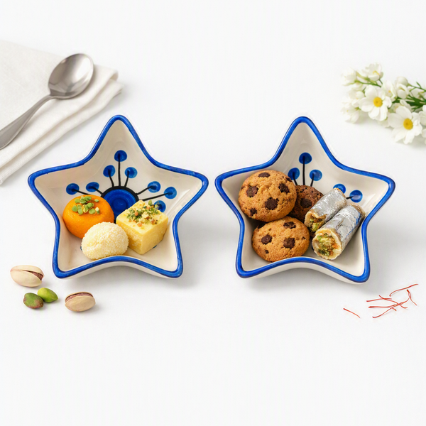 Two star-shaped ceramic serving dishes with blue floral borders, one containing cookies and chocolate-covered rolls, and the other containing colorful sweets, styled on a white surface with a spoon, pistachios, and small white flowers.