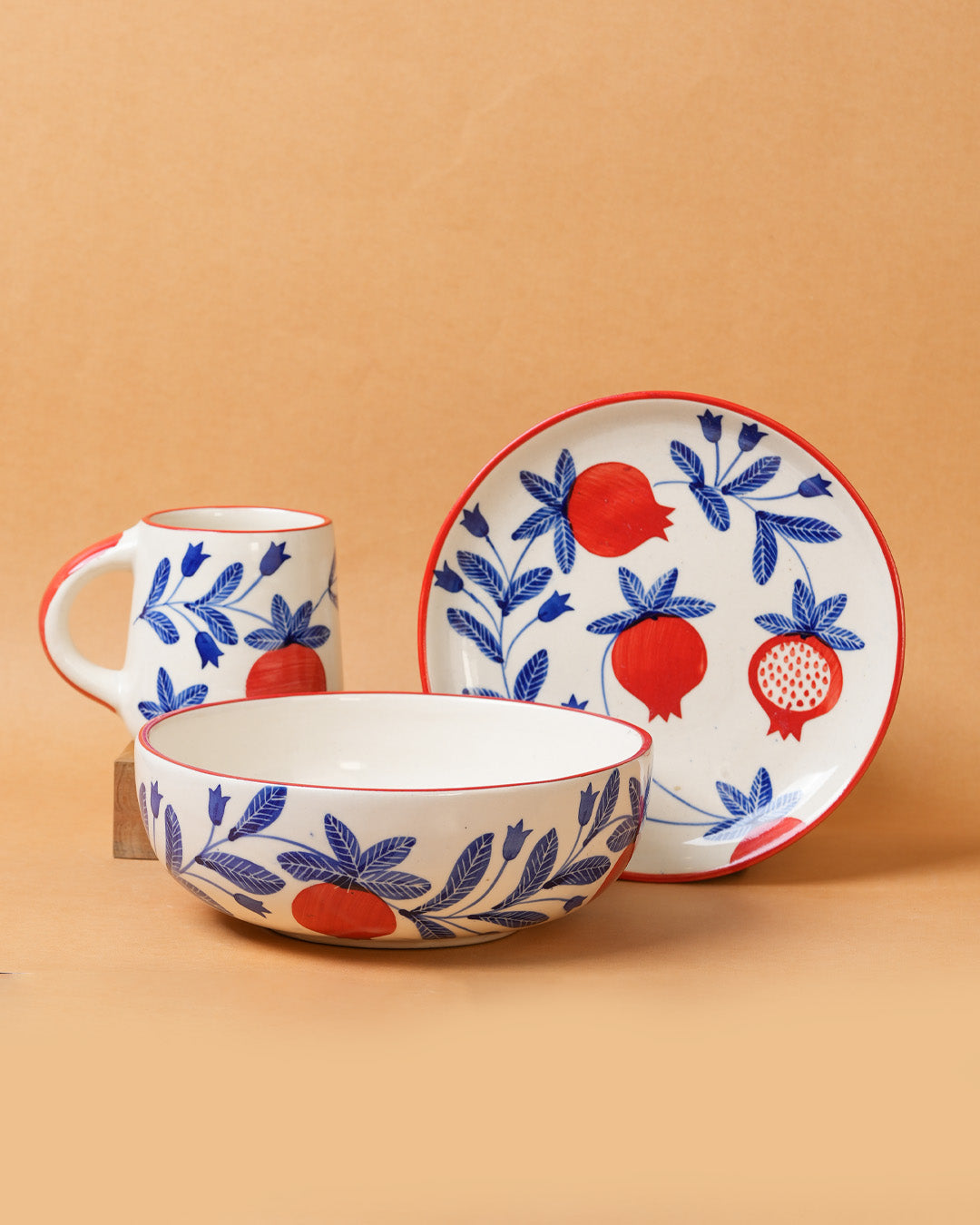 Ceramic set with bowl, mug, and plate featuring red pomegranate and blue leaf design on a beige background