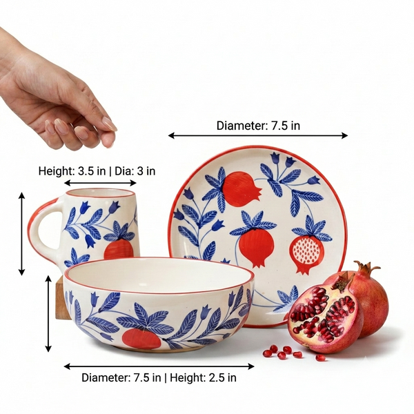 A set of ceramic tableware with a blue and red pomegranate pattern, shown with dimension arrows and labels indicating diameter and height. A hand is visible at the top left for scale, with pomegranate props on the right against a white background.