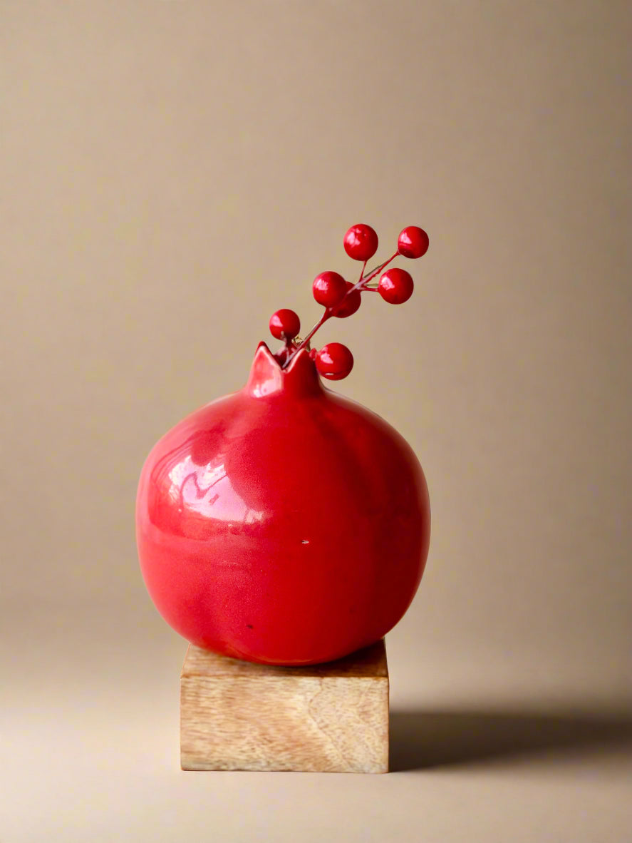A red ceramic vase with a shape resembling a pomegranate, placed on a wooden stand, with a decorative element that resembles a branch with leaves or berries extending from the top.