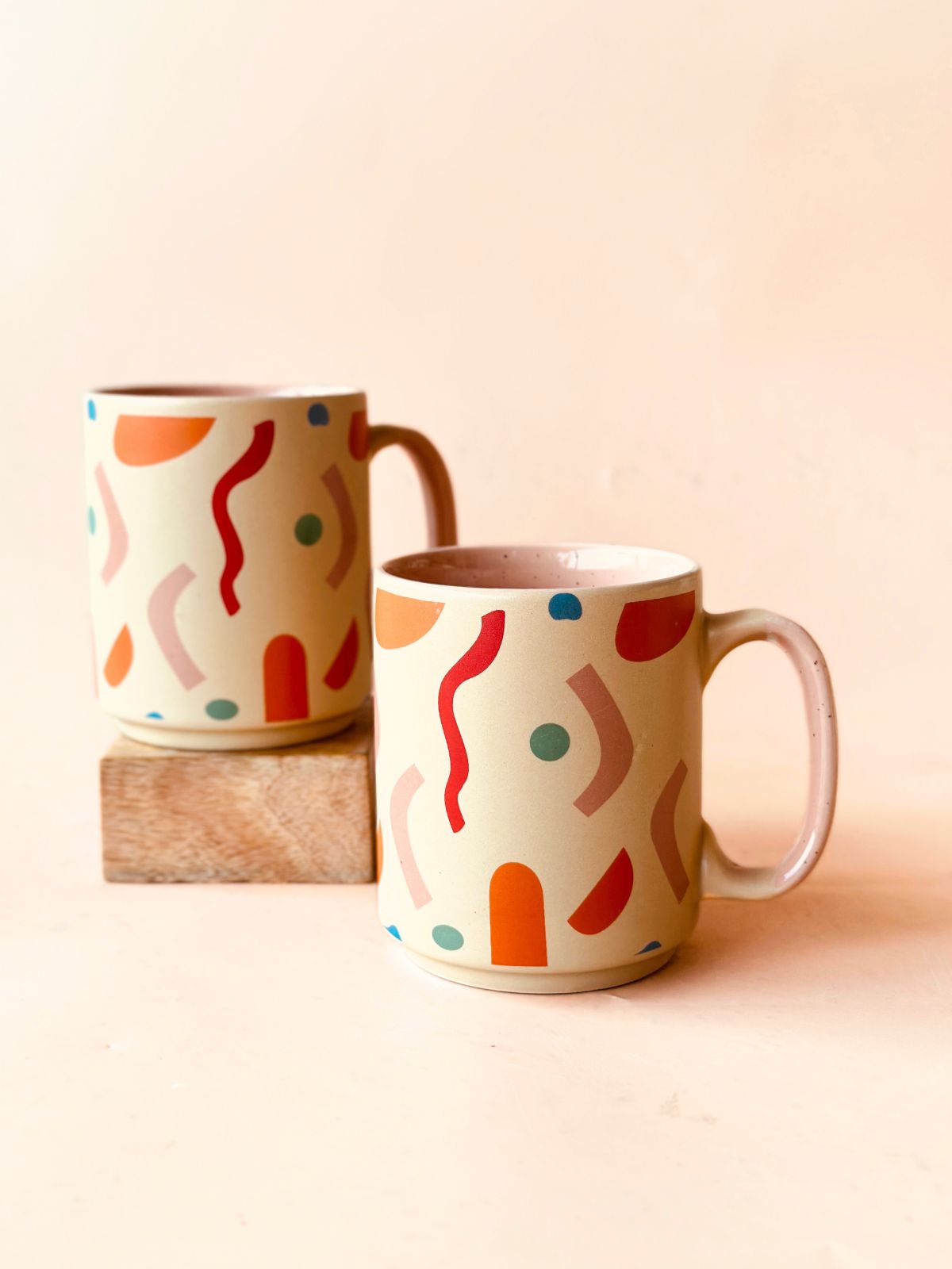 Two ceramic stoneware mugs with a colorful geometric confetti pattern on a beige background, displayed on a wooden surface.