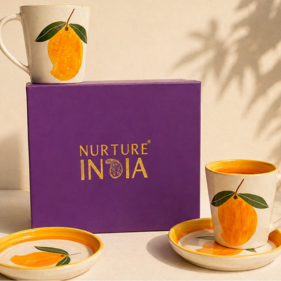 Nurture India ceramic mug and saucer set with a matching box. The white mugs and saucers feature a yellow-orange mango illustration with green leaves, set against a warm beige background with leaf shadows.