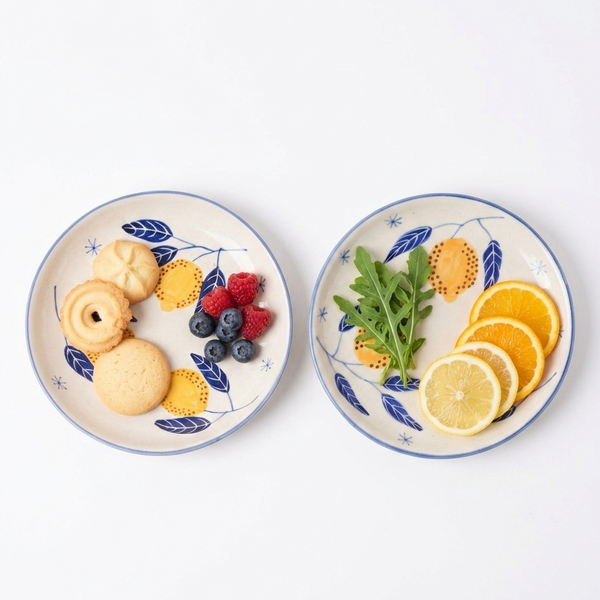 Two ceramic plates with decorative blue leaf patterns, one containing cookies and berries, the other with fruits and greens.