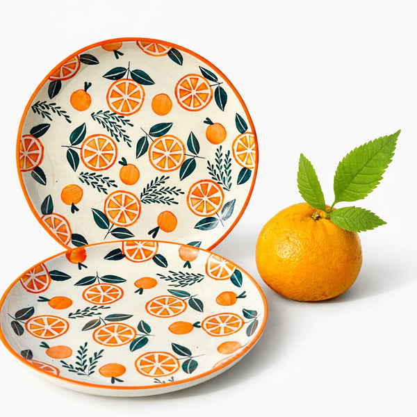 Two ceramic plates with orange and leaf pattern next to a real orange on a white background