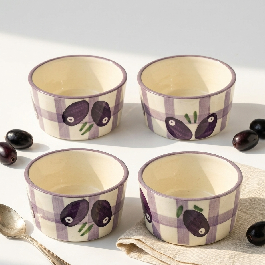 Four small ceramic bowls arranged in a 2x2 grid on a white surface, decorated with a purple and white plaid pattern and dark purple olive illustrations. A silver spoon and folded beige cloth are visible, with a few black olives scattered around.