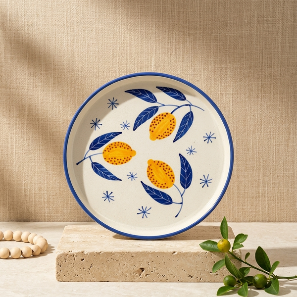 A round ceramic plate with a blue rim and a hand-painted lemon and leaf pattern, displayed on a stone block with a wooden bead garland and green leaves in the foreground against a textured beige backdrop.