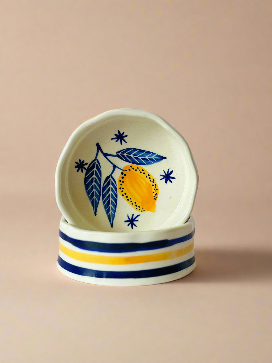 A set of two ceramic cereal bowls with handpainted leaf and lemon designs in colors blue, green, and yellow on a white background with blue striped patterns on the outside.