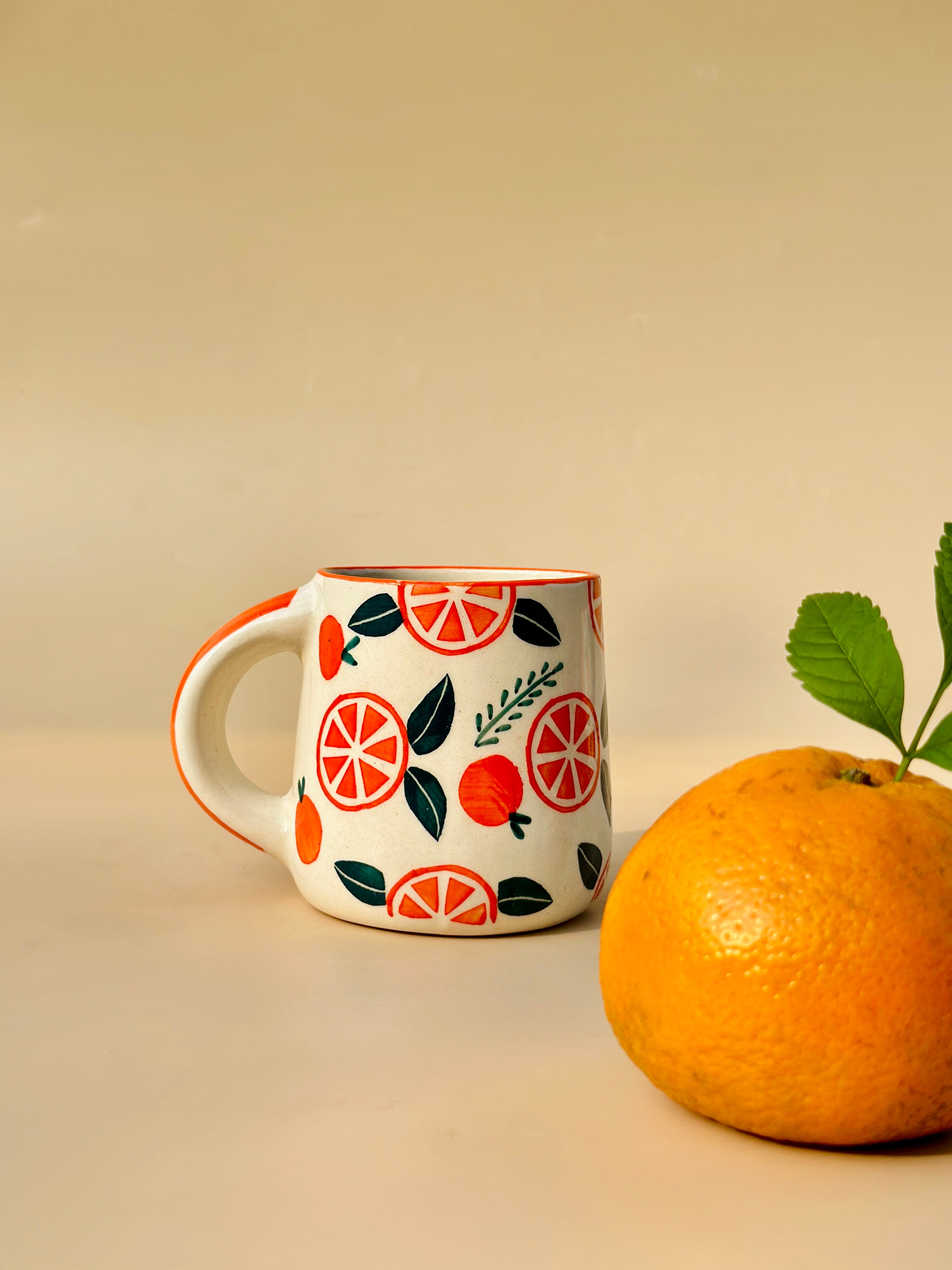 A ceramic mug with a lavender hue and a pattern of orange slices and green leaves, accompanied by a fresh orange fruit with a leaf on it, against a beige background.