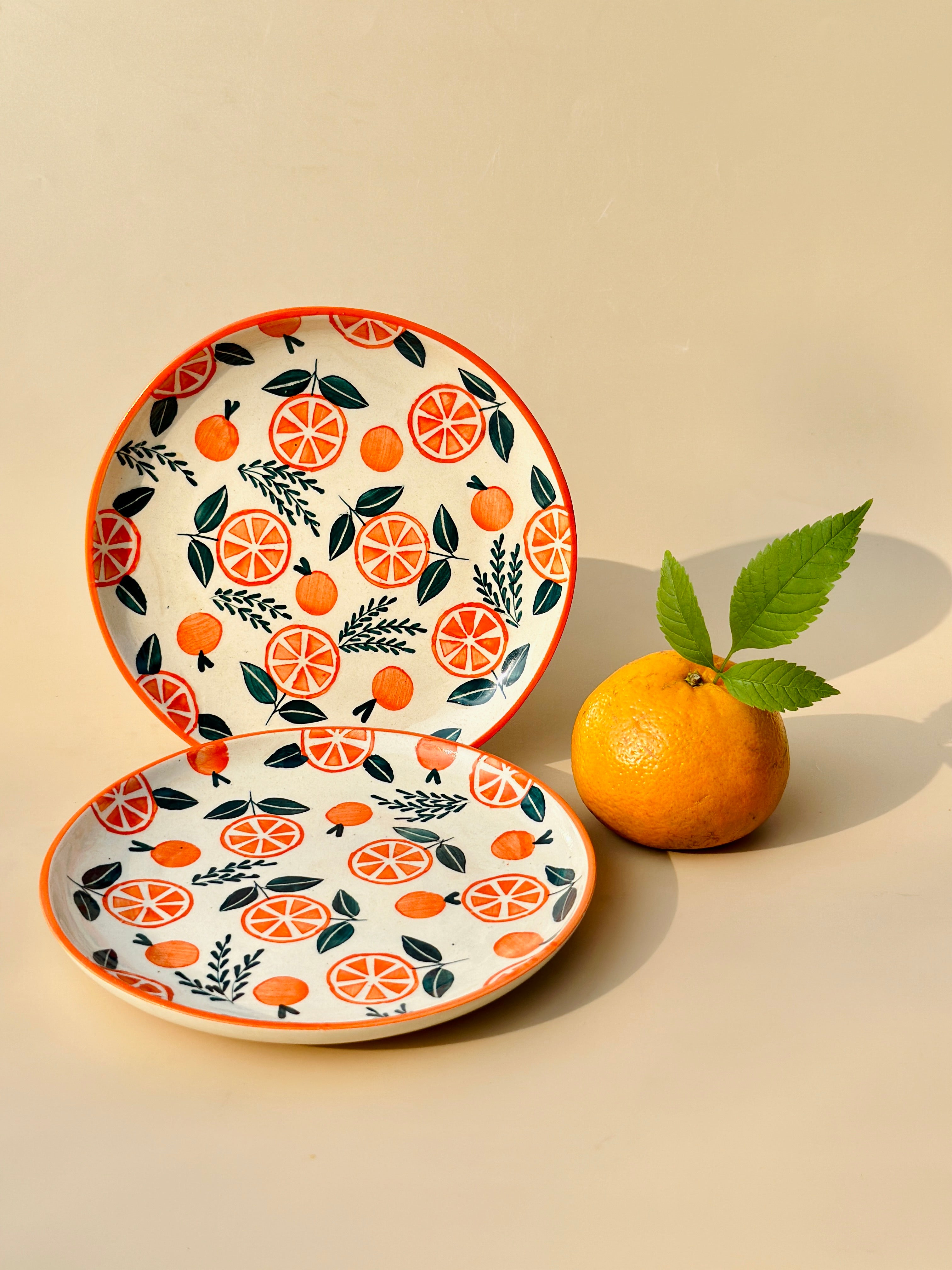 Two ceramic plates with a orange and green floral pattern, alongside a fresh orange with a leaf on it, all placed on a beige surface.