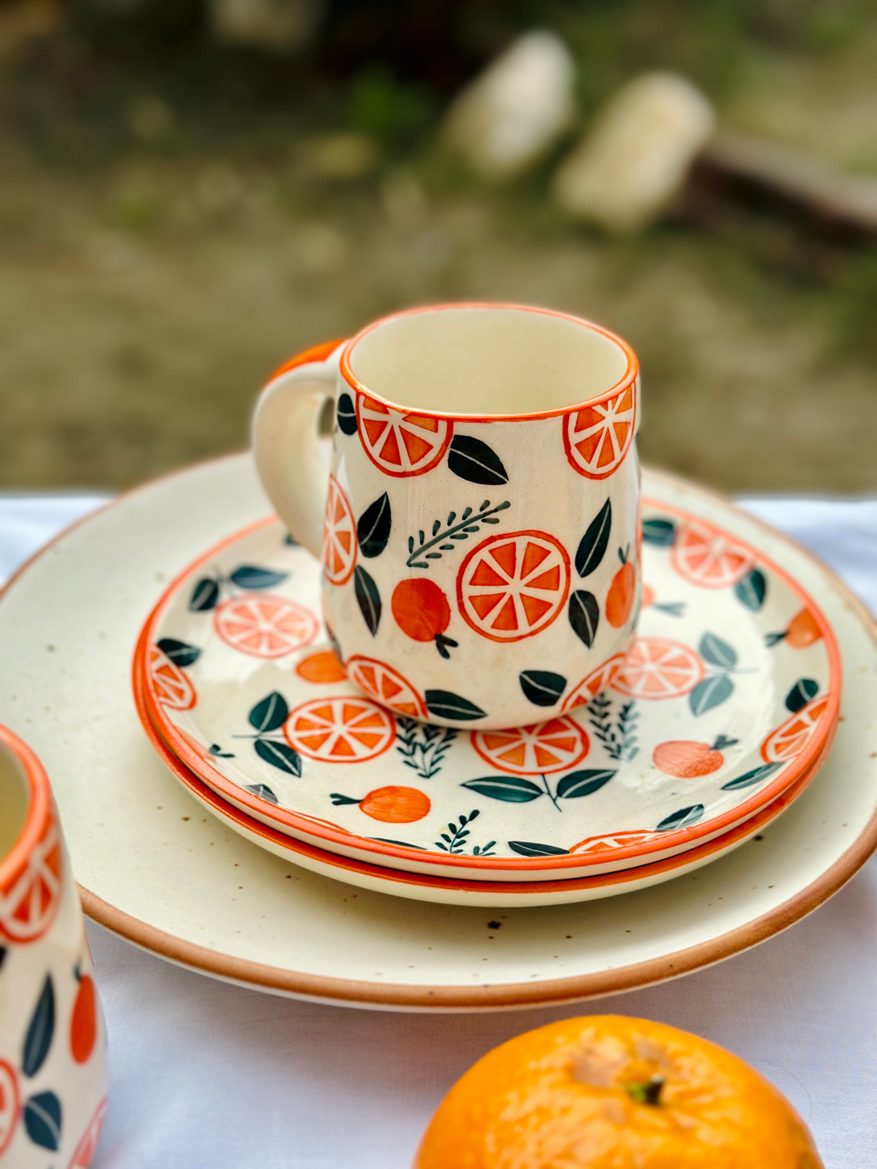 A ceramic mug and plate set with orange color and floral patterns, displayed with a ripe orange beside them.