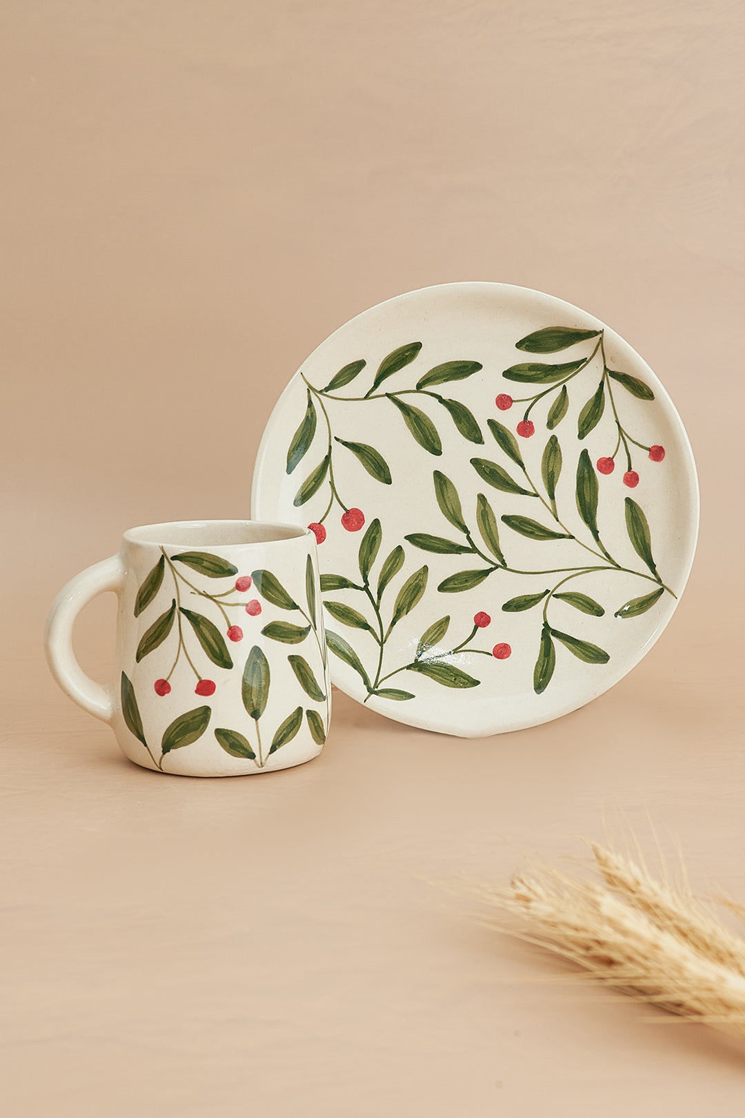 A ceramic plate and mug set with a green and red olive leaf pattern on a beige background, displayed on a table with a wheat stem in the corner.