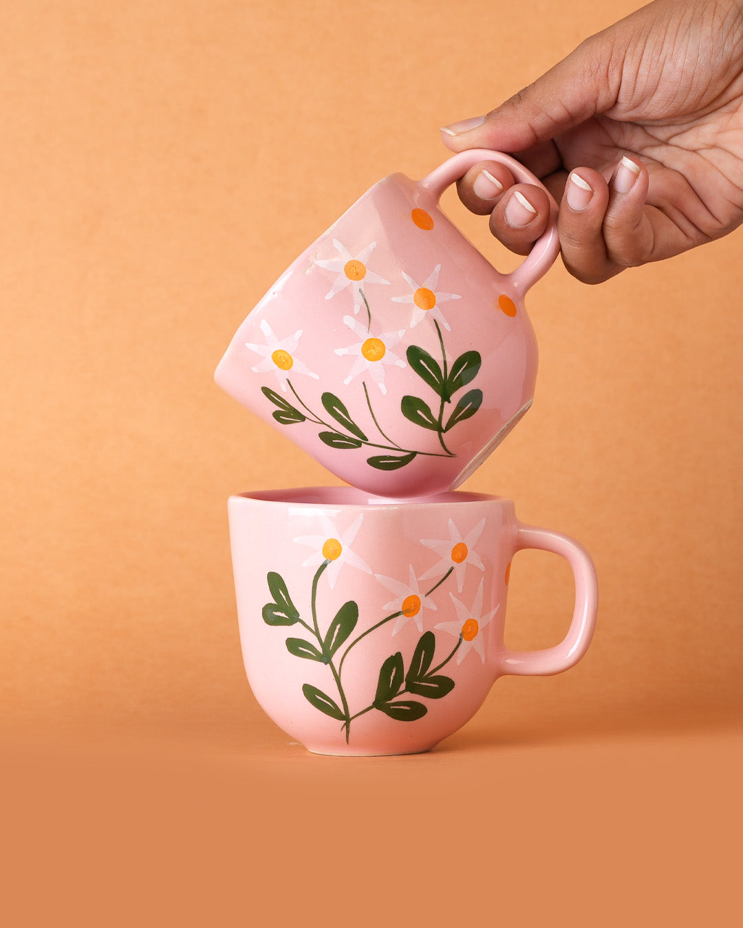 Pink mugs with floral designs held by a hand against an orange background