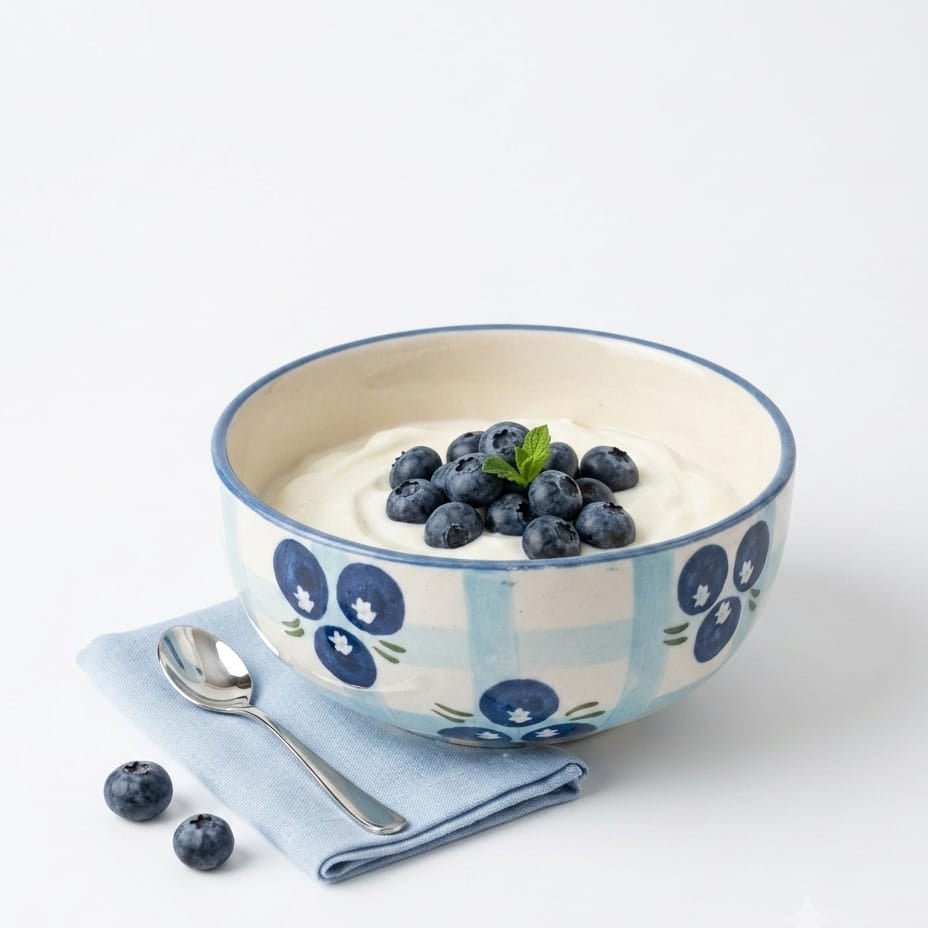 A ceramic bowl with a blue and white gingham pattern and blue floral motifs, filled with yogurt and topped with fresh blueberries and a mint leaf, styled on a white background with a blue napkin and a spoon.