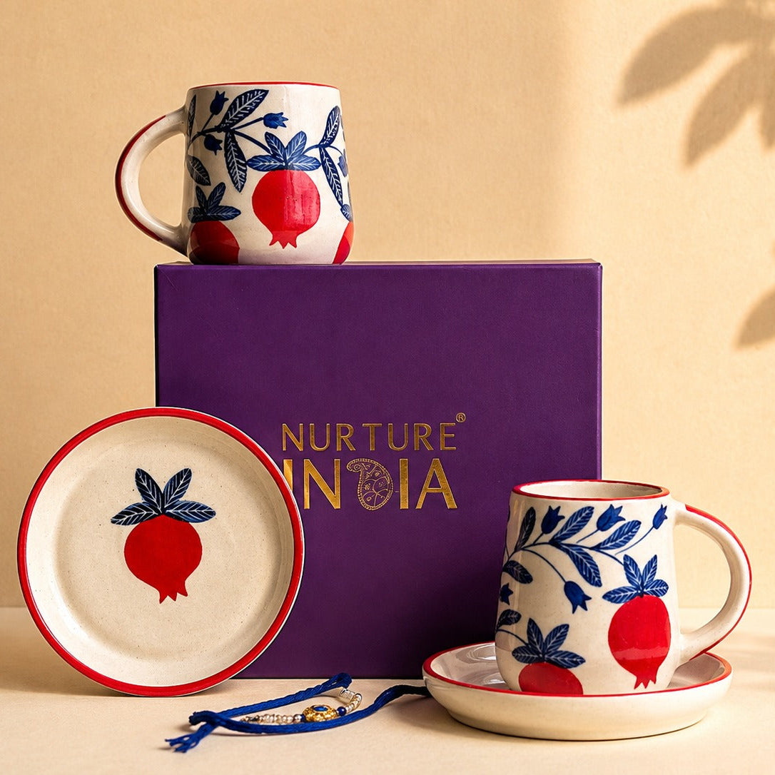 A ceramic mug, matching saucer, and round plate with a red and blue pomegranate motif, displayed alongside a purple Nurture India gift box on a beige surface with leaf shadows.