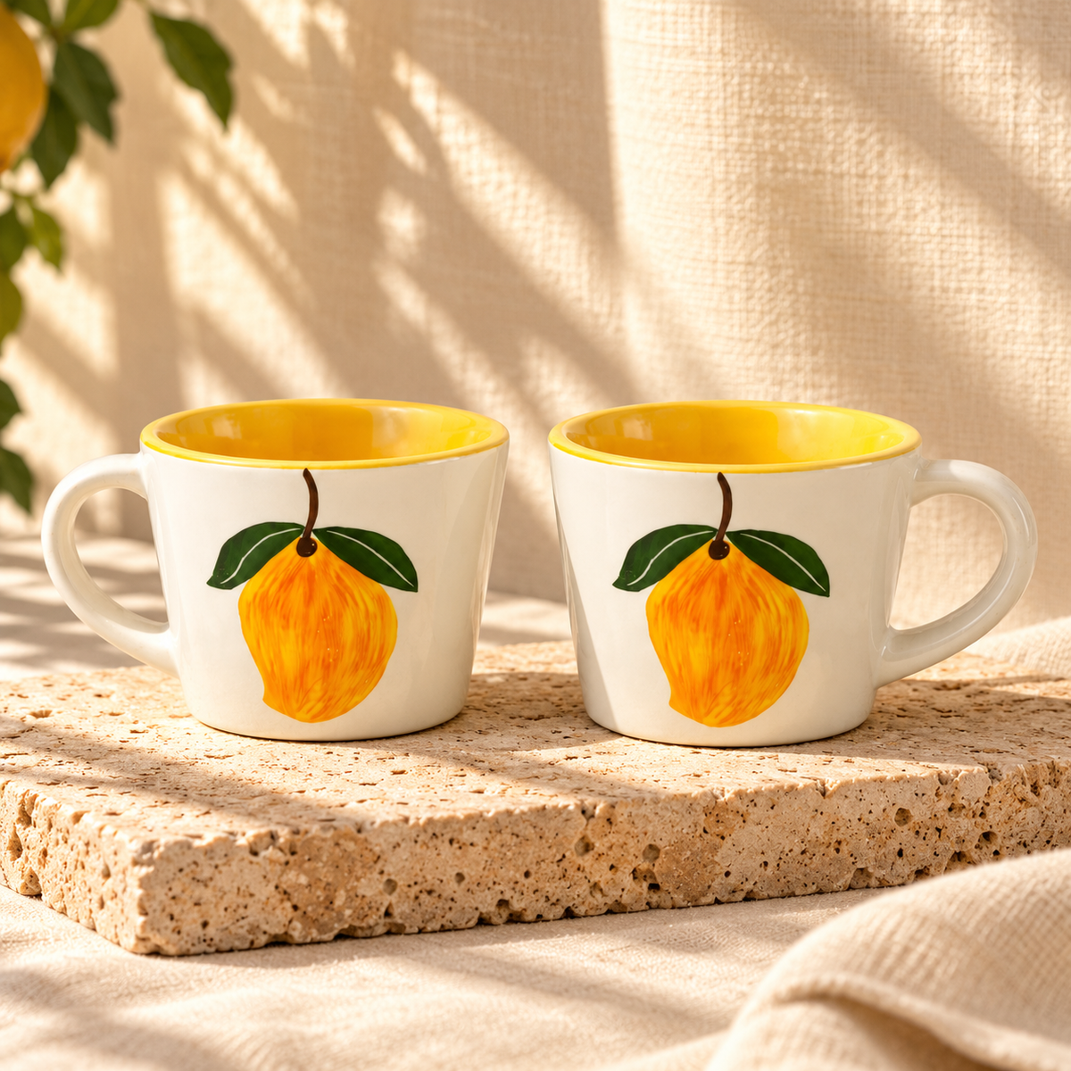 Two white ceramic mugs with yellow interiors and a painted lemon design on the front, displayed on a textured stone slab against a warm beige fabric backdrop with soft leaf shadows.