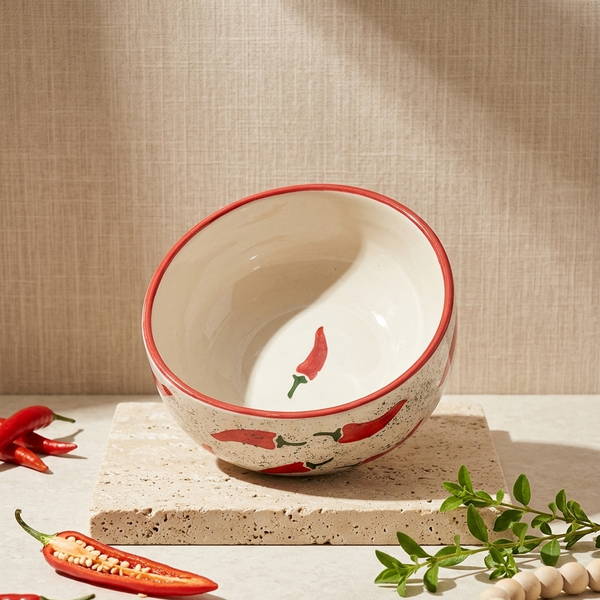 A ceramic bowl with a red rim and chili pepper illustrations, displayed on a stone slab with red chili peppers and green leaves in a styled setting.