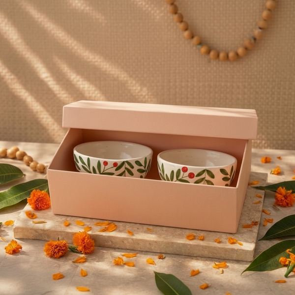 Two small white ceramic bowls with green leaf and red berry patterns, presented inside an open blush pink box on a stone surface with orange marigold flowers and green leaves around them.