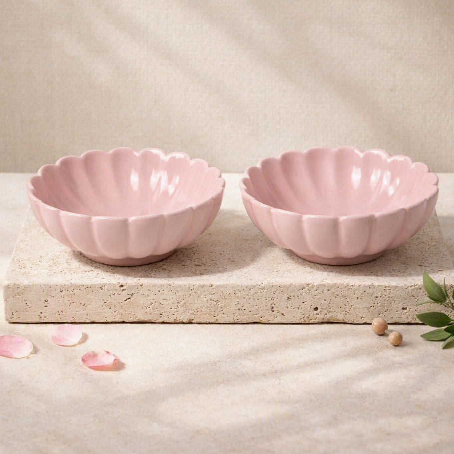 Two pink scalloped ceramic bowls displayed side by side on a textured stone slab, with soft shadows and decorative petals on a neutral beige background.